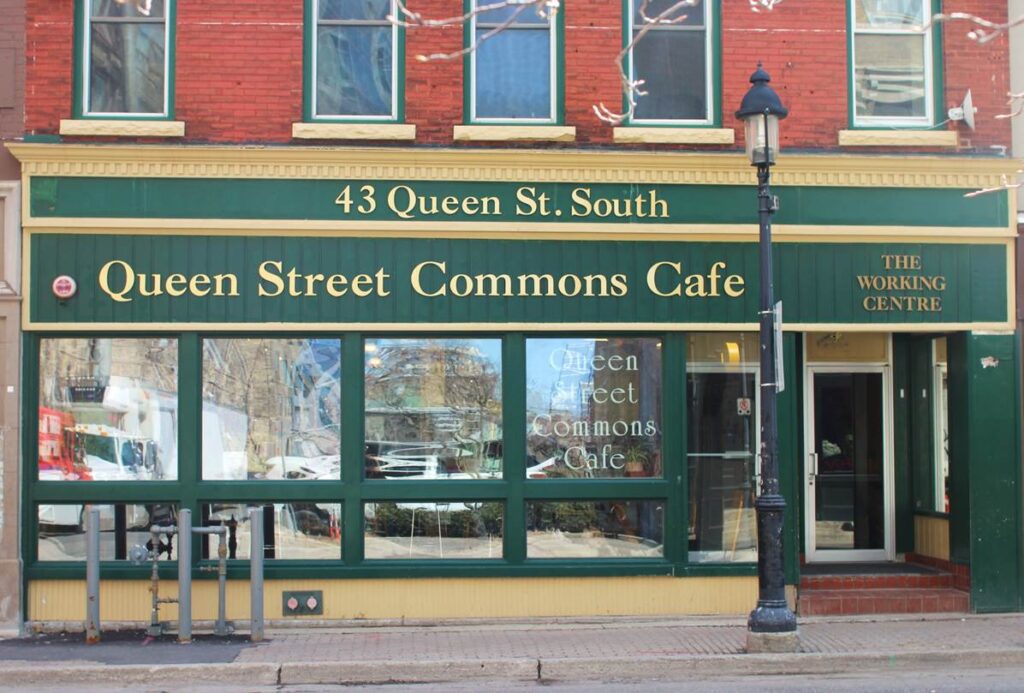 The street level of the 43 Queen Street South building has green signage and window framing with tan coloured trim. A sign with large letters reads Queen Street Commons Cafe.