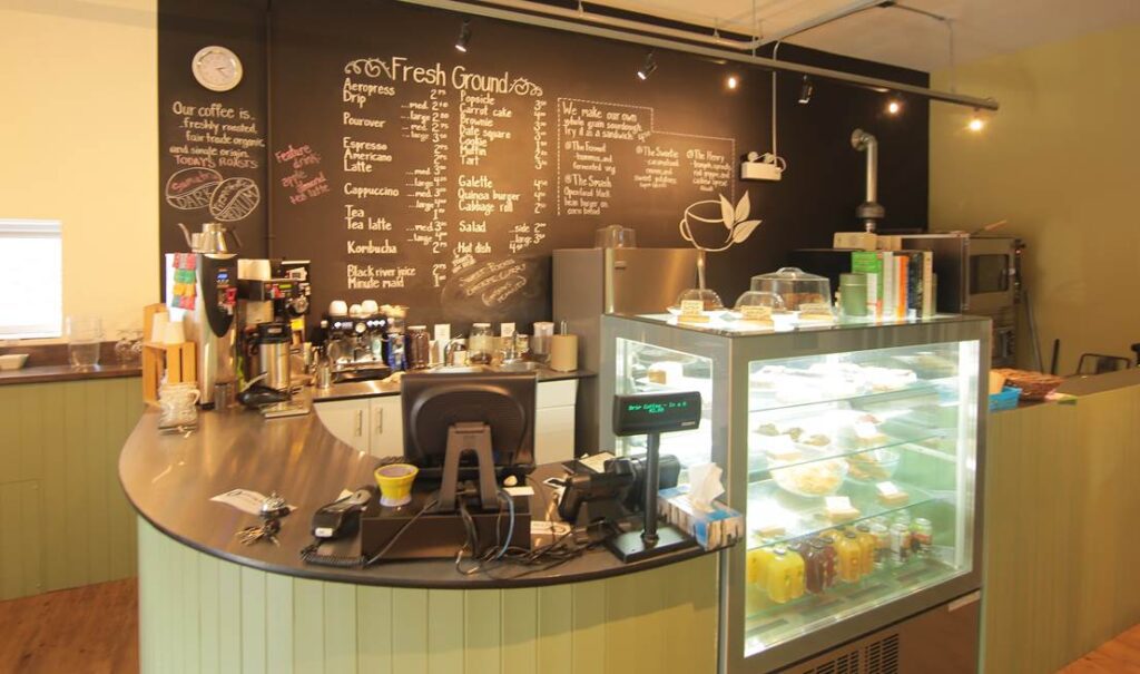 Retail counter at Fresh Ground Cafe with a display case showing food and drinks. Menu items are written in chalk on a large blackboard wall in the background.