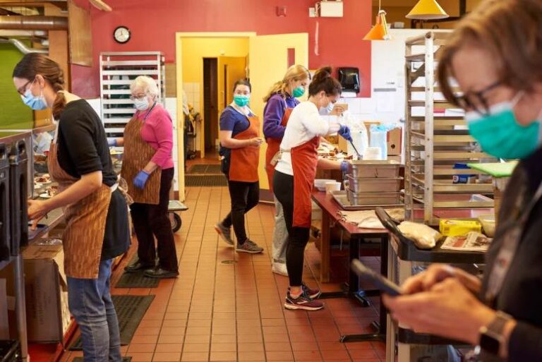 Several Saint John Kitchen's workers wearing aprons and face masks stand in the kitchen preparing food and plates. A woman in the foreground checks her phone.