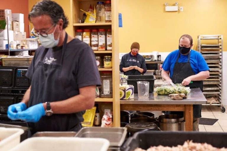 Three workers wearing black aprons and face masks prep food inside Queen Street commissary kitchen.