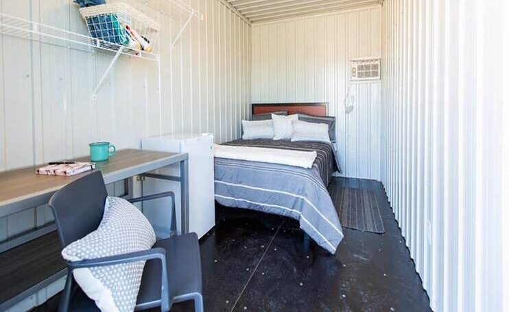 A bed with pillows, a chair and desk, and a small fridge furnish the inside of a cabin at Erbs Road Shelter