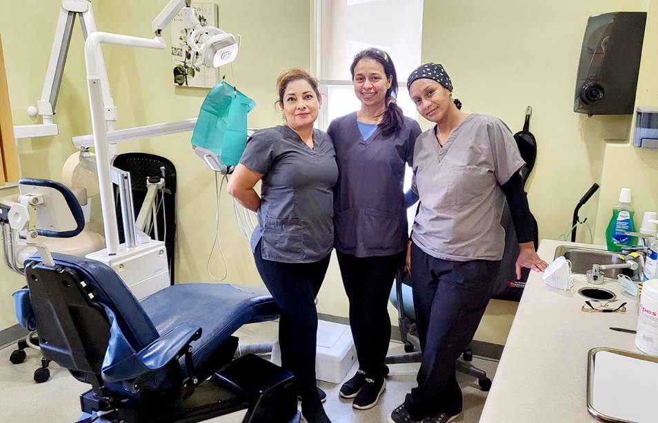 Three staff members of Community Dental pose for a photo in room of dental equipment and a dental procedure chair